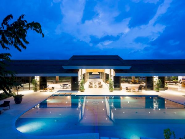 The image shows a luxurious modern building with a large swimming pool, illuminated beautifully against a blue evening sky.