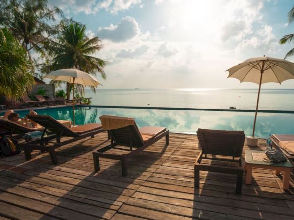 A serene beachfront scene with lounge chairs and umbrellas on a wooden deck, overlooking a calm, sunlit sea and palm trees.