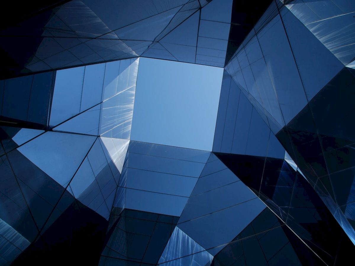 The image shows a geometric, abstract view of a building with glass and mirrored surfaces reflecting the sky, creating a kaleidoscopic effect.