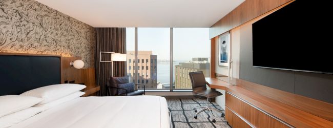 The image shows a modern hotel room with a large bed, TV, desk area, and a window with a city view.