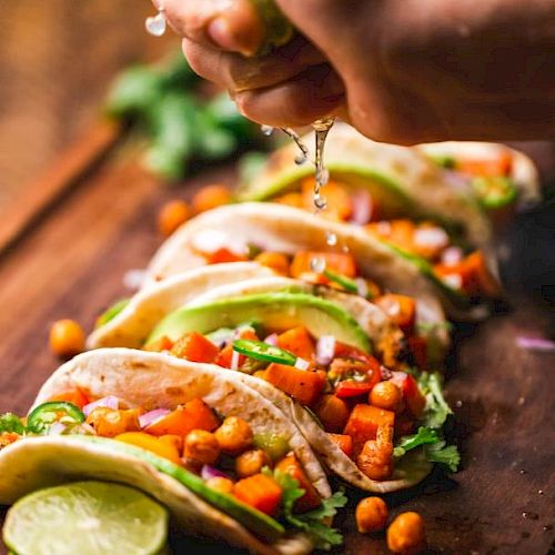 A hand squeezing lime over tacos filled with chickpeas, avocado, and vegetables on a wooden board.