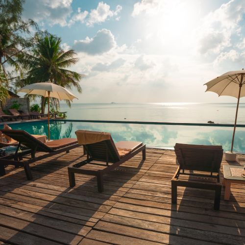 A serene beach scene with lounge chairs, umbrellas, and a pool overlooking the ocean under a bright sky with some clouds.