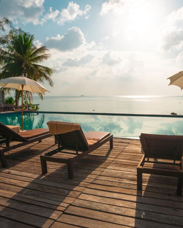 A serene beach scene with lounge chairs, umbrellas, and a pool overlooking the ocean under a bright sky with some clouds.