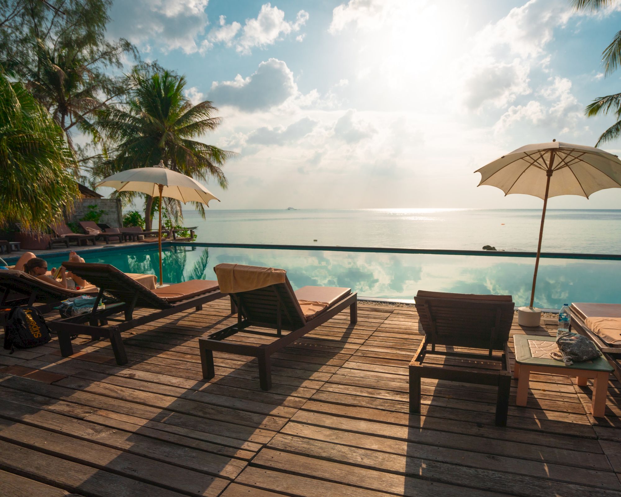 Poolside scene with lounge chairs, umbrellas, palm trees, and a view of a tranquil ocean under a partly cloudy sky.