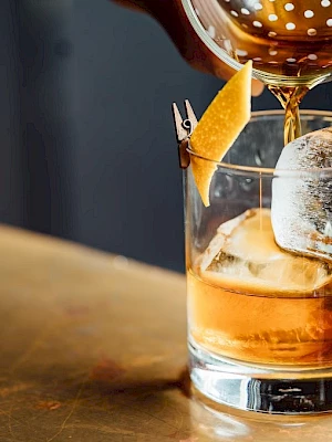 A cocktail is being poured into a glass with ice and a lemon twist on a bar counter.