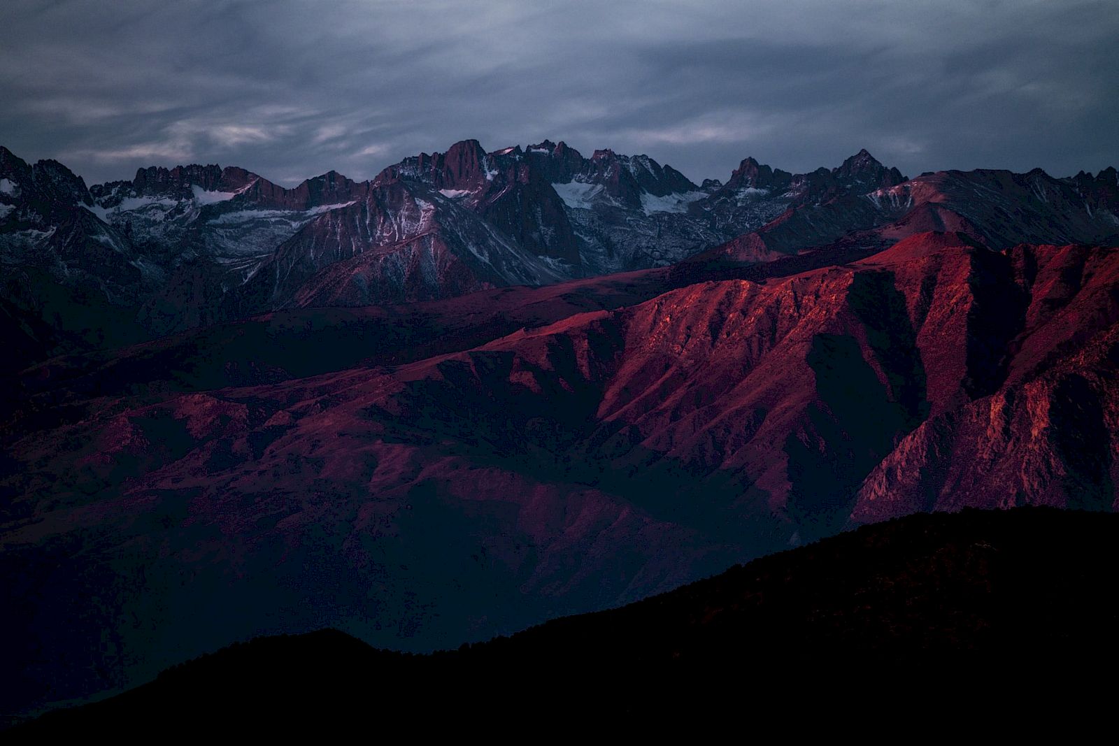 A dramatic mountain landscape at dusk, with rugged peaks and red-tinted slopes under a moody, clouded sky.