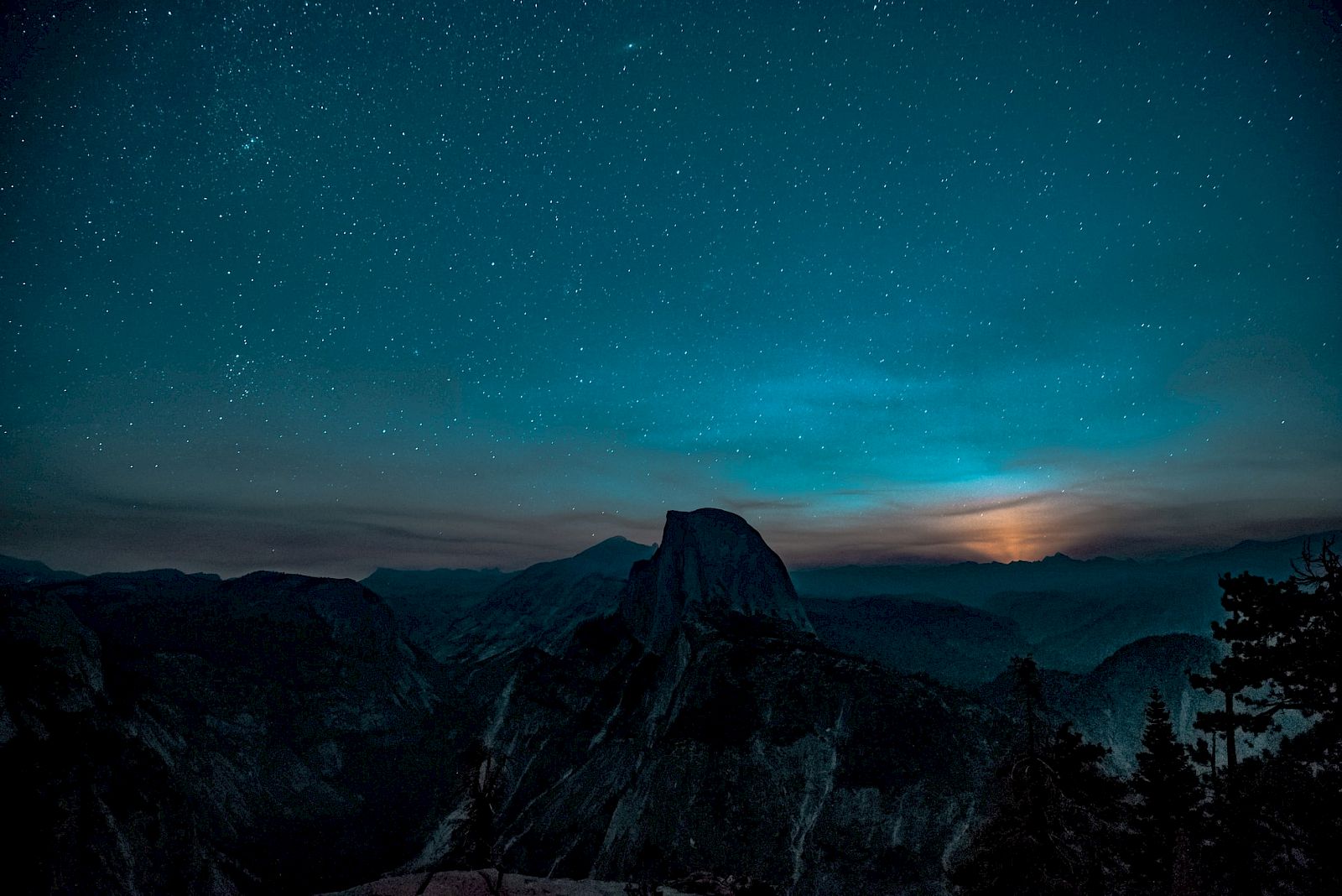 A starry night sky over a mountainous landscape with a silhouette of a prominent peak and faint glow on the horizon.