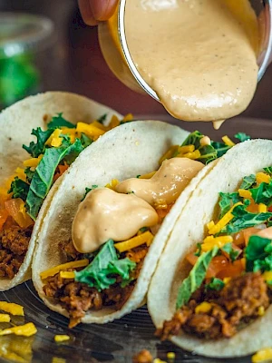 Three tacos filled with ground meat, cheese, and greens, with a sauce being poured over them on a plate.