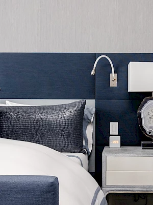 A modern bedroom scene features a bed with blue accents and a white nightstand holding a lamp and decor against a neutral wall.