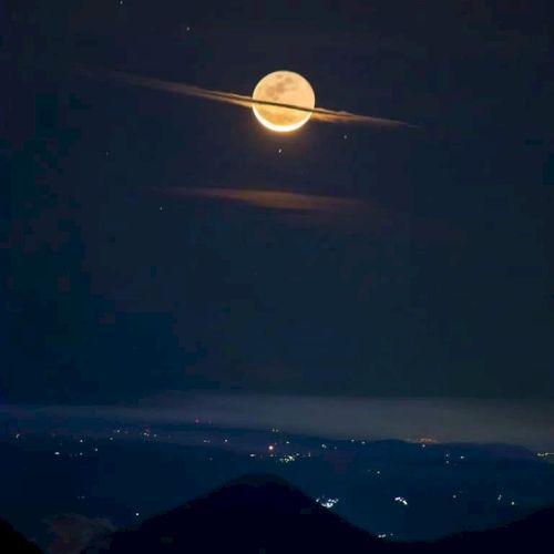 A full moon appears to be wearing a cloud "saturn-like" ring above a landscape with distant city lights in the darkness.