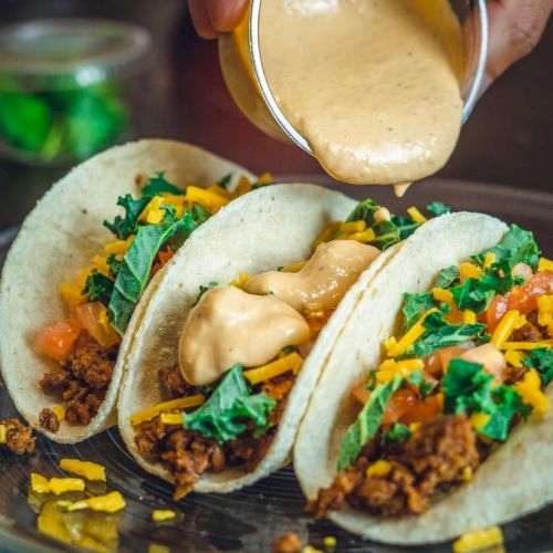 Three tacos filled with ground meat, cheese, and greens are being topped with a creamy sauce on a plate.