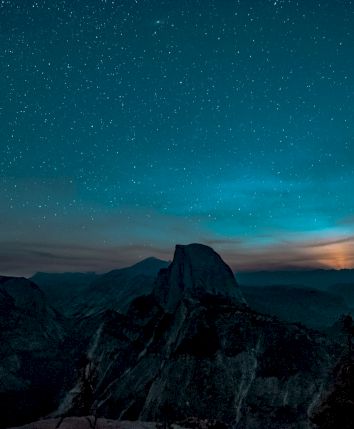 A starry night sky over a mountainous landscape with a distinct peak and trees silhouetted against the horizon.