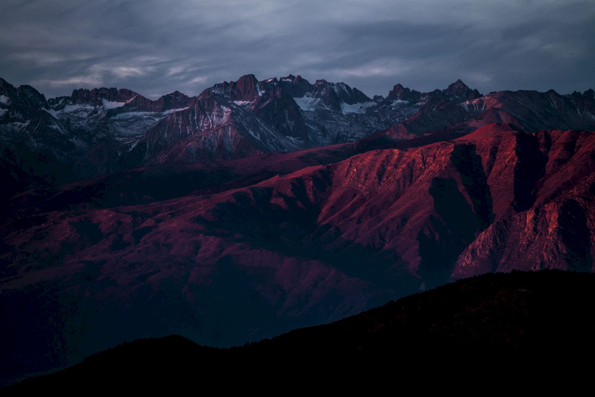 A mountain range is visible under dark, cloudy skies, with red sunlight casting vibrant hues across the peaks and ridges.
