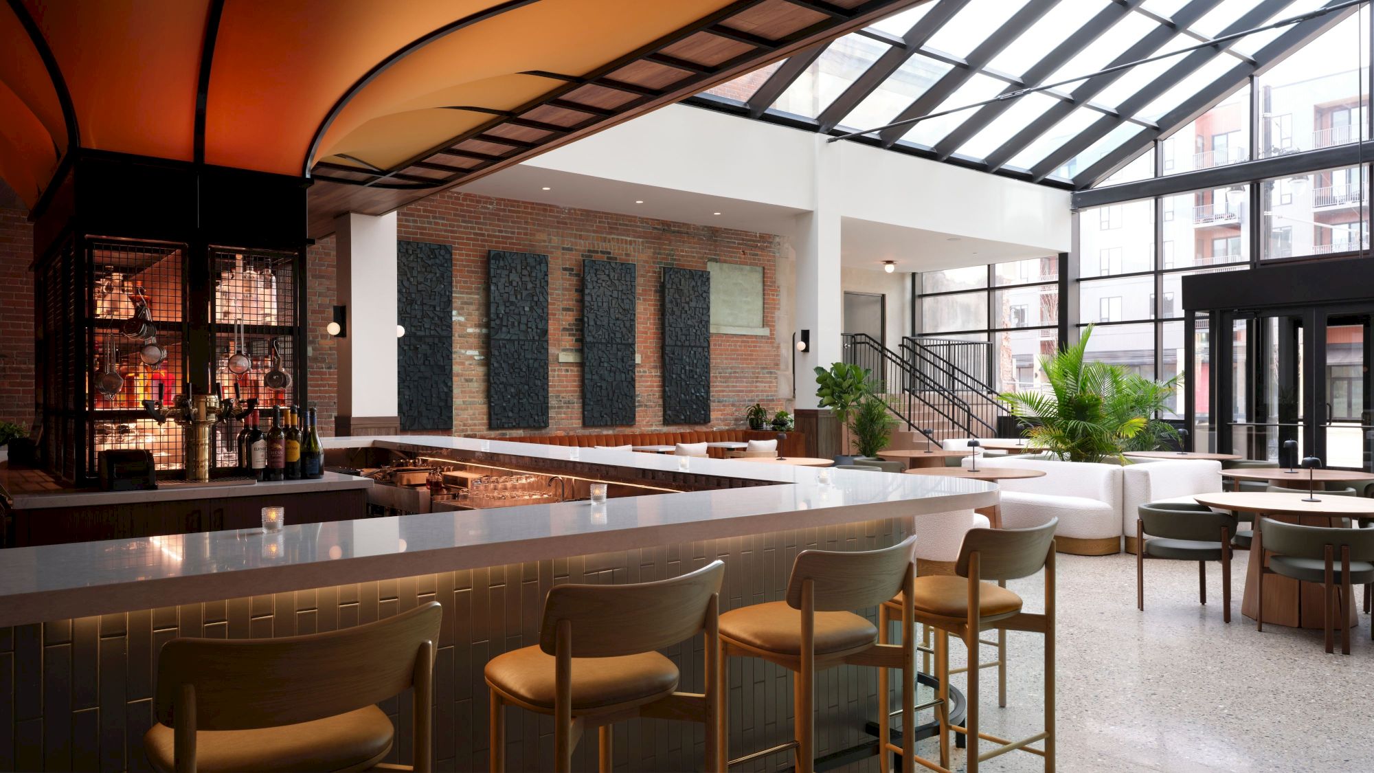 A modern bar with high chairs, a counter, cozy seating, large windows, and a stylish design featuring brick walls and indoor plants.