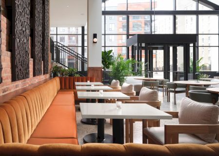 The image shows a modern restaurant with orange booth seating, white tables, and large windows letting in natural light, creating an airy feel.