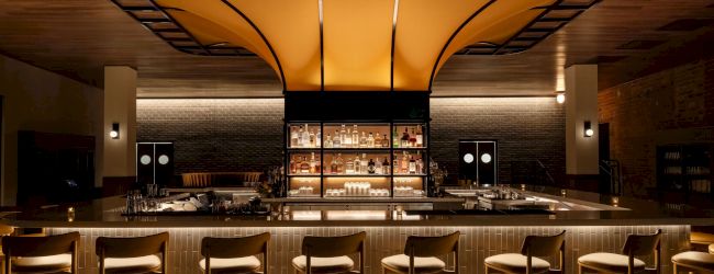 The image shows a stylish bar with wooden stools, a well-lit counter, and a striking overhead canopy design.