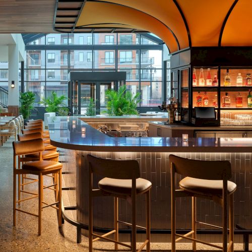 A modern bar with a curved counter, high stools, and a backlit shelf of bottles, set in a warmly lit space with large windows.