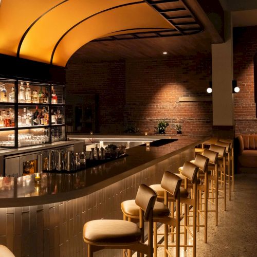 This image shows a stylish bar with a curved counter, bar stools, a liquor display, and a modern, cozy atmosphere.