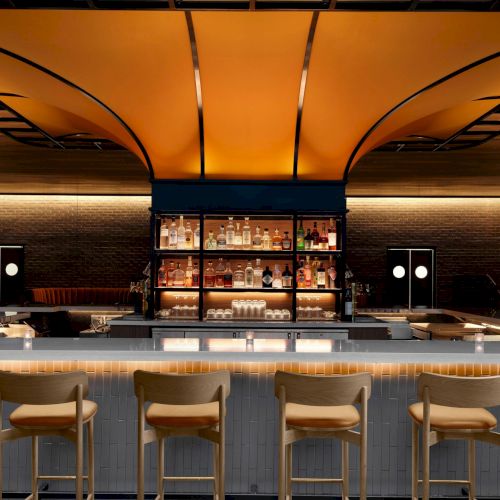A stylish bar interior with a curved ceiling, well-stocked shelves, and a row of wooden chairs along the counter, warmly lit.
