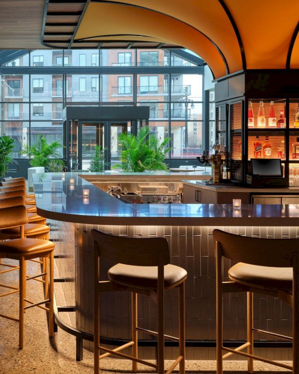 The image shows a modern bar with a curved counter, high chairs, and a well-stocked shelf, featuring a cozy and stylish interior.