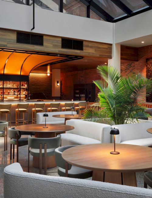 The image shows a modern, stylish lounge with a bar, round wooden tables, green plants, and warm lighting, creating a cozy atmosphere.