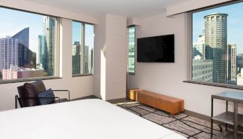 A modern hotel room with large windows showcasing a city view, furnishings include a bed, chair, wall-mounted TV, and a wooden bench.