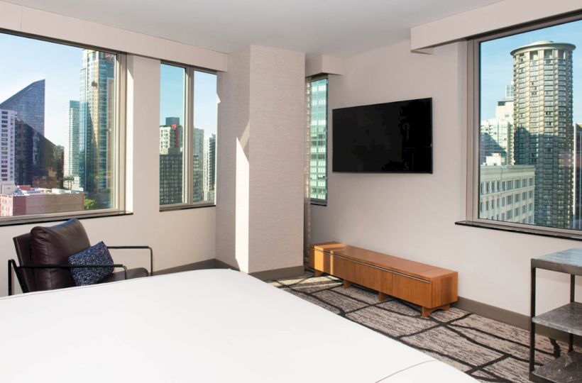 A modern hotel room with large windows showcasing a city view, furnishings include a bed, chair, wall-mounted TV, and a wooden bench.