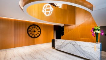 The image shows a modern lobby with a wooden spiral ceiling, marble reception desk, large clock, and decorative lights and flowers.