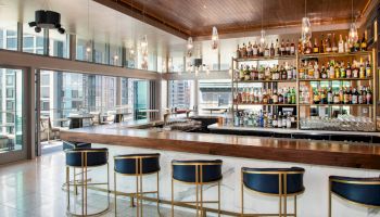 A modern bar with stylish stools, a well-stocked liquor shelf, and large windows overlooking a cityscape.