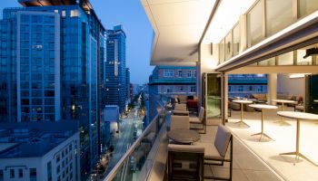 A modern cityscape view from a stylish balcony at dusk, featuring high-rise buildings and outdoor seating with warm lighting.