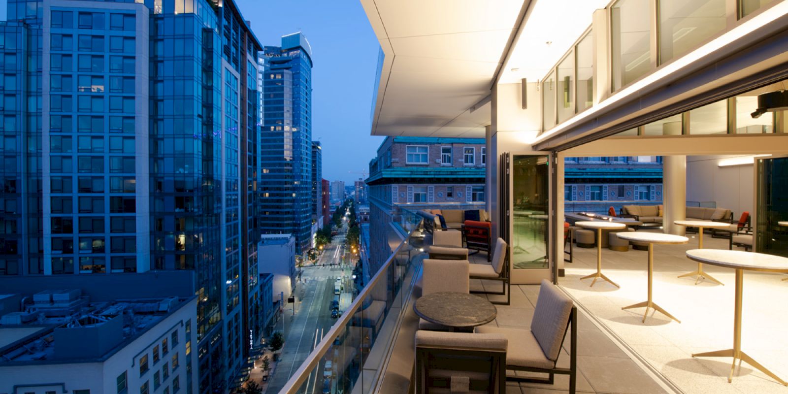 Rooftop view with modern seating area, overlooking a city street lined with tall glass buildings, captured during twilight.