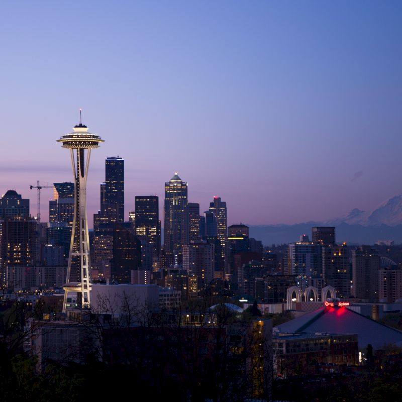 A city skyline during twilight with a prominent tower and a mountain in the background against a purple sky.