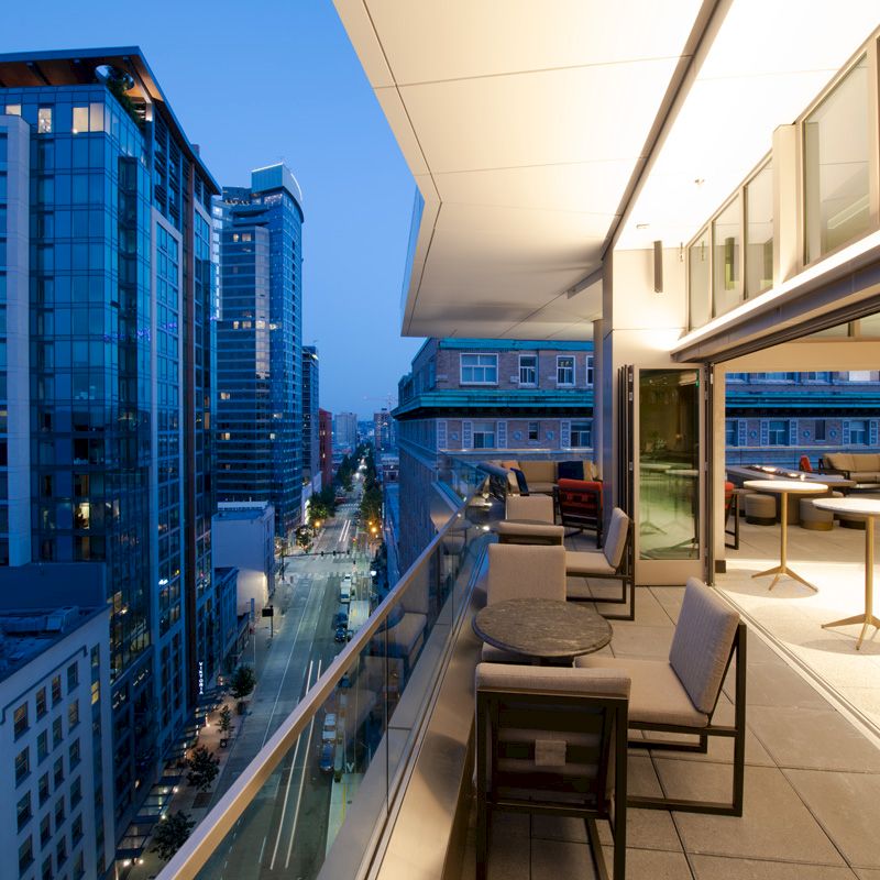 A modern rooftop terrace with seating overlooks a cityscape filled with tall buildings under a twilight sky.