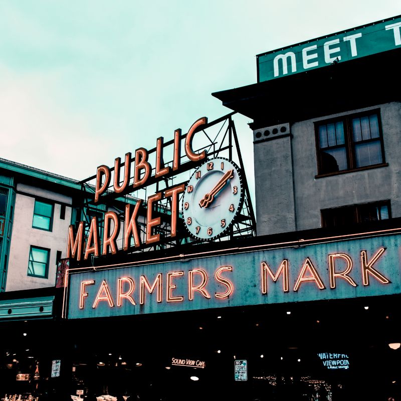 It's an image of a public farmers market sign with a clock above, in an urban area with adjacent buildings visible.