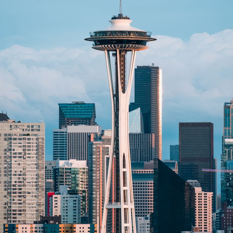 The image shows the Space Needle with a backdrop of skyscrapers in Seattle, capturing a cloudy blue sky above the cityscape.