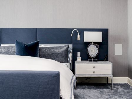 A modern bedroom with a blue upholstered bed, white nightstand, sleek lamp, and decorative pillow, all on a soft carpeted floor.
