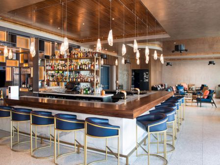 The image shows a modern bar with a counter, bar stools, liquor shelves, and pendant lights in an open, stylish setting.