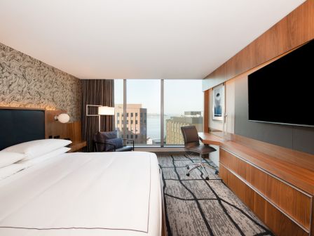 A modern hotel room features a large bed, a desk, a chair, and a flat-screen TV, with a view of buildings through a large window.