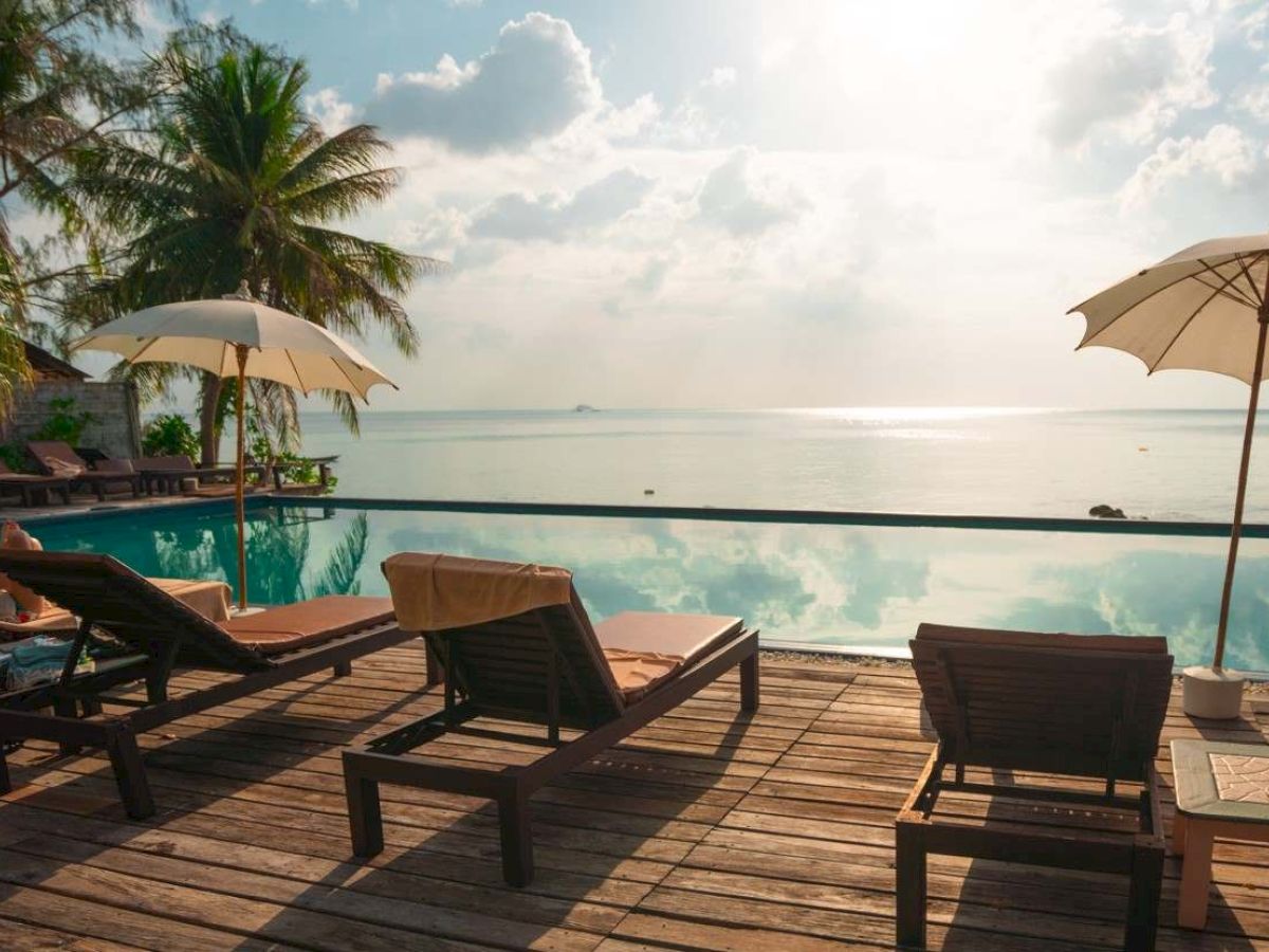 Sunlit beach scene with loungers and umbrellas on a wooden deck by a pool, overlooking a calm sea and palm trees in the background.