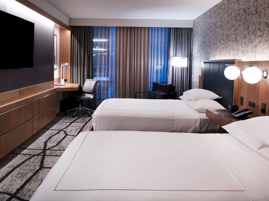 The image shows a modern hotel room with two beds, a large TV, a work desk, and contemporary lighting, featuring a stylish carpet and decor.
