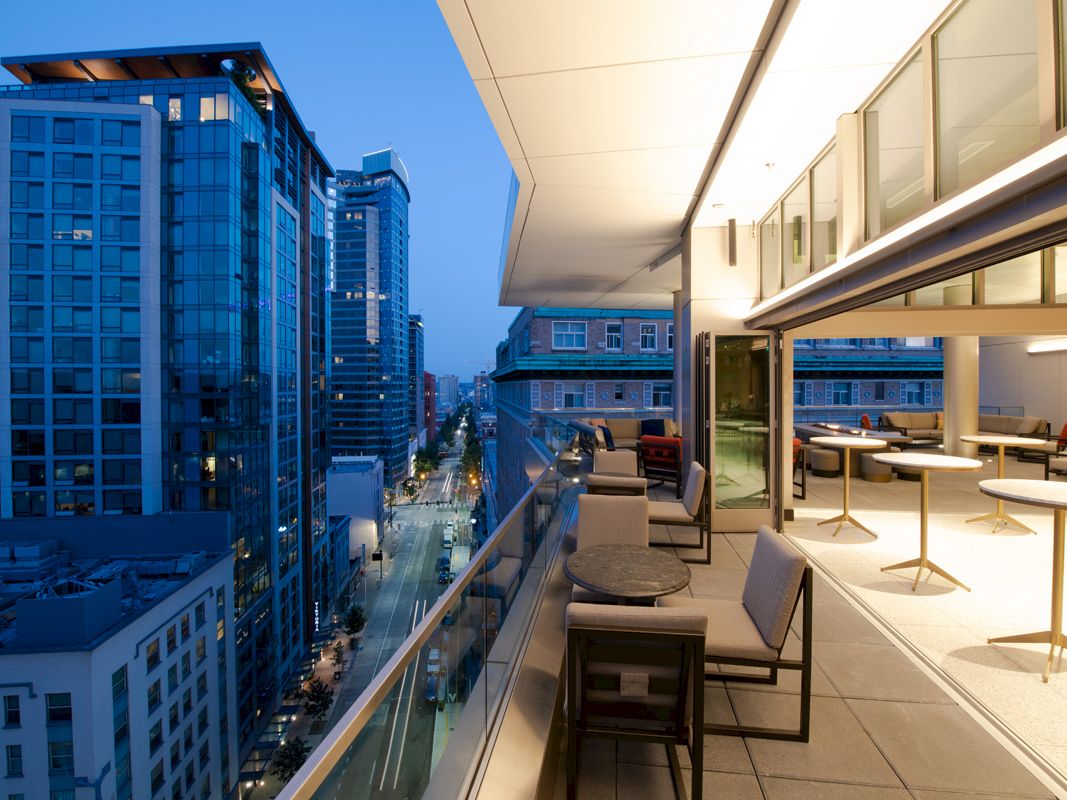 A modern rooftop terrace overlooking a cityscape at dusk, featuring seating areas with tables and lounge chairs under ambient lighting.