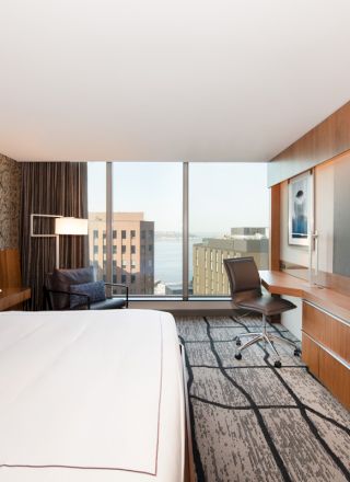 A modern hotel room features a large bed, flat-screen TV, desk, and window view of city buildings, with elegant decor and lighting.