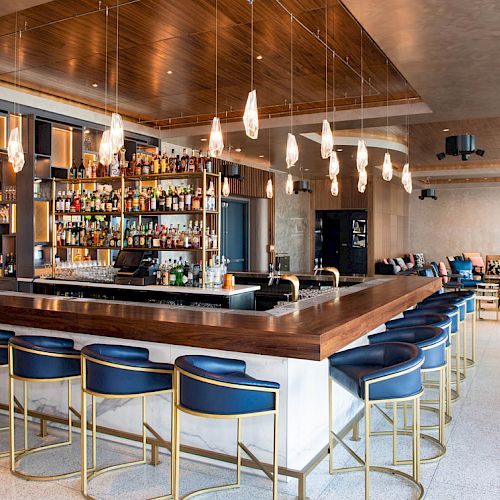 The image shows a modern bar with blue stools, hanging lights, and a stocked counter. There’s seating in the background and a stylish interior.