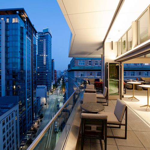 A modern rooftop patio with seating overlooks a city street and tall buildings illuminated by evening lights.