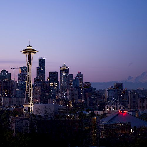 A city skyline at dusk featuring a distinctive tower, with mountains visible in the background, under a clear sky.
