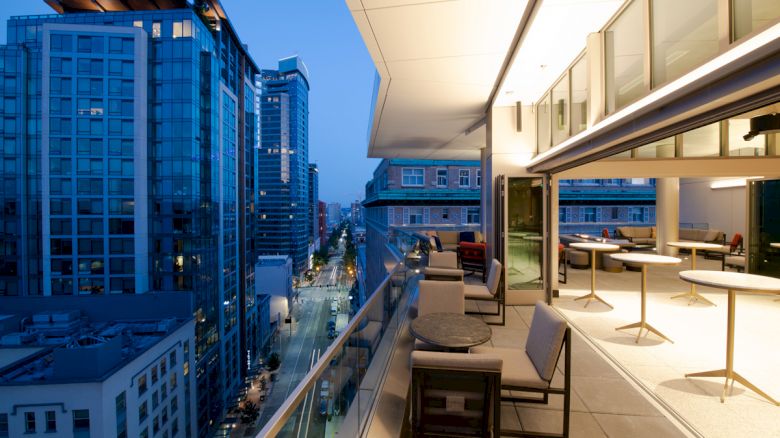 A modern rooftop terrace with seating areas overlooks a cityscape of high-rise buildings at dusk.