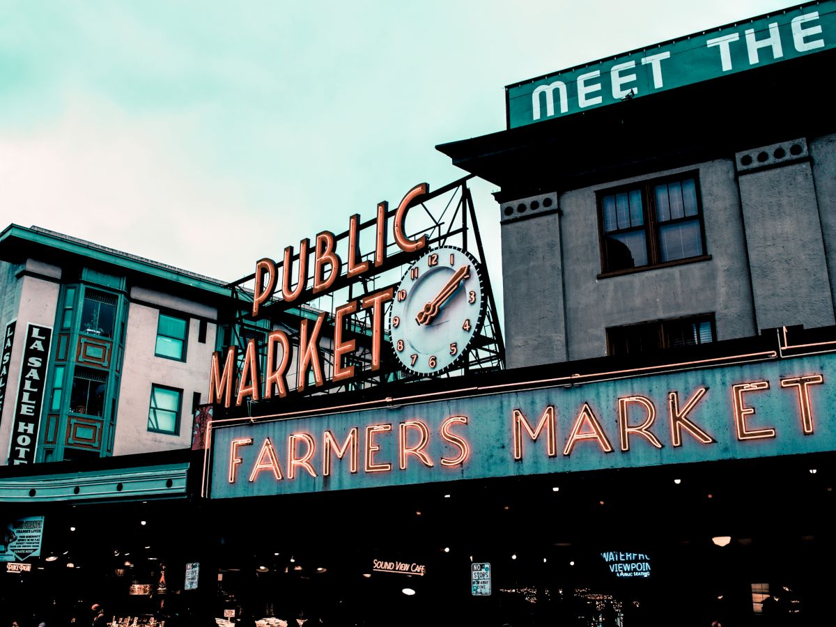 The image shows the iconic entrance signs of a public farmers market with neon lights and a clock, with buildings in the background.