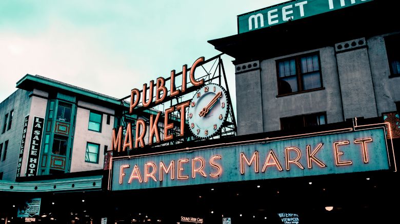 The image shows the iconic entrance signs of a public farmers market with neon lights and a clock, with buildings in the background.