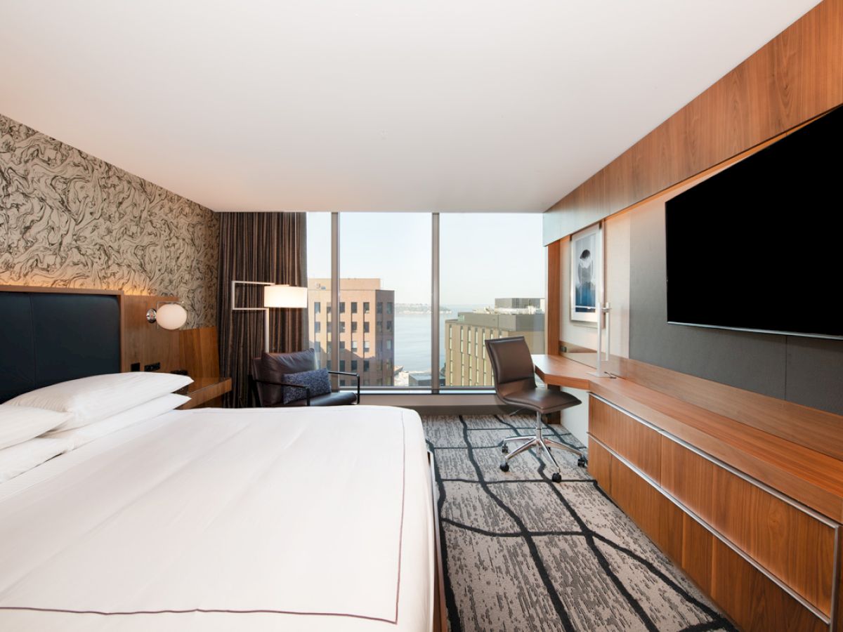 A modern hotel room with a large bed, wall-mounted TV, desk, and city view through a large window, featuring sleek wooden furnishings.