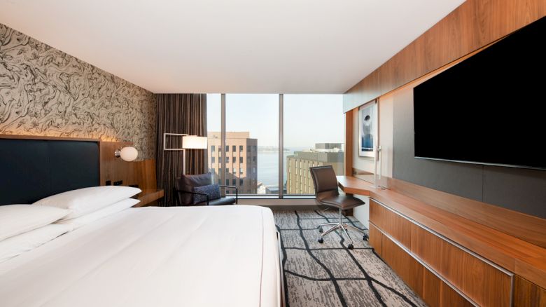 A modern hotel room with a large bed, wall-mounted TV, desk, and city view through a large window, featuring sleek wooden furnishings.